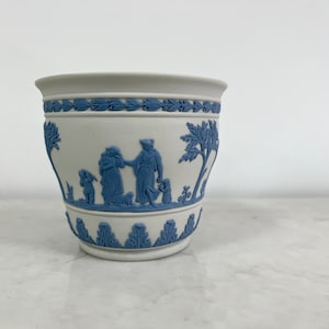 Może przedstawiać: Biała ceramiczna doniczka z niebiesko-białym wzorem przedstawiającym scenę z ludźmi i drzewem. Doniczka ma zaokrąglone dno i szeroki rant.