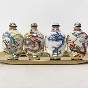 May include: Four antique Chinese snuff bottles with detailed hand-painted designs of dragons and phoenixes in vibrant colours. The bottles are displayed on a gold-coloured oval tray, showcasing their intricate artwork and cultural significance.