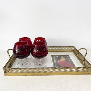 May include: A rectangular brass serving tray with a decorative lattice border and two side handles. Inside the tray are four ruby red glass snifters with clear stems and a small rectangular print featuring a portrait of a person in red clothing.