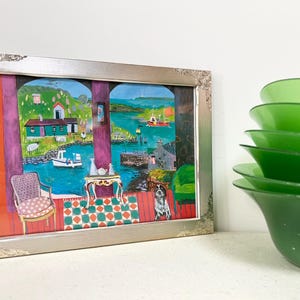 May include: A framed painting with a silver ornate frame depicts a coastal scene with boats, houses, and a dog. The painting is set against a white background, with a stack of green bowls to the right.