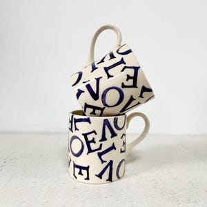 May include: Two white ceramic mugs with blue lettering that says "LOVE" stacked on top of each other.