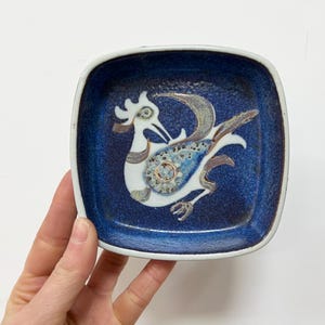May include: Square ceramic dish with a deep blue speckled background and a white border. The dish features a stylized bird design in white, blue, and brown. A decorative piece for serving or display.