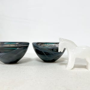 May include: Two dark gray glass bowls with a swirling pattern and a white horse figurine. The bowls have a glossy finish and a rounded shape. The horse is carved from a translucent material.