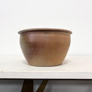 Può includere: Un vaso rotondo in ceramica marrone con un bordo largo. Il vaso ha una forma leggermente affusolata e una palette di colori naturali e terrosi. Un piccolo segno è visibile sul lato. Adatto per uso interno o esterno.