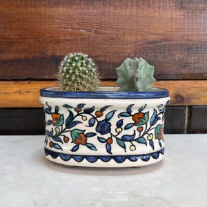 May include: Oval ceramic planter with a white background and blue, green, orange, and yellow floral pattern. The planter is filled with two small green cacti.