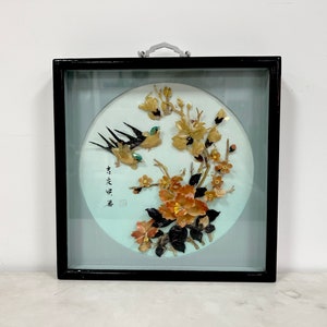 May include: A framed wall art piece featuring a round, light blue background with a floral design made of various colored stones. Two birds are depicted in flight, with black wings and yellow bodies. The frame is black wood with a silver hanging hook.