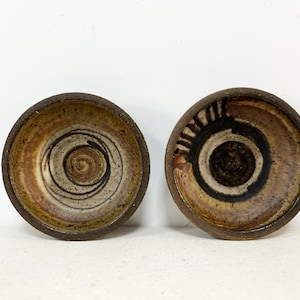 May include: Two handmade ceramic bowls with a rustic aesthetic. One bowl features a spiral design in brown and black, while the other has a concentric circle pattern. The bowls have a dark brown rim and a light brown interior.