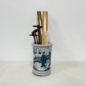 Może przedstawiać: Cylindryczny ceramiczny pojemnik z niebiesko-białym wzorem inspirowanym Azją, zawierający linijki, pędzle i inne narzędzia. Pojemnik ma około 13 cm wysokości i 8 cm średnicy.