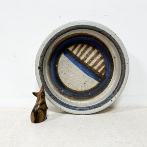 May include: A handmade ceramic bowl with a speckled, off-white exterior and a blue and brown interior design. A small, carved wooden figurine is next to the bowl. The bowl's diameter is approximately 8 inches.