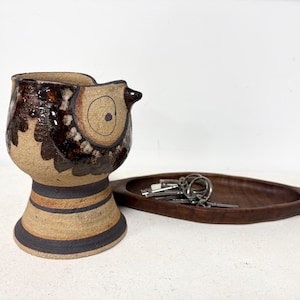 May include: A ceramic owl-shaped container with brown and tan tones, featuring a detailed eye design. Beside it, a dark wooden, leaf-shaped tray holds a set of keys. The items are set against a white background.