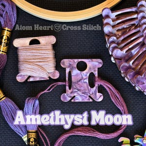 May include: A flat lay featuring cross-stitch supplies. Includes spools of thread, a wooden embroidery hoop, and a purple thread organizer. The text "Amethyst Moon" is displayed in a purple font. The brand name "Atom Heart Cross Stitch" is also visible.