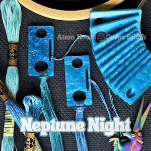 May include: An overhead shot features cross-stitch supplies. Several blue thread bobbins and skeins are arranged with blue acrylic thread organizers. The text "Neptune Night" is displayed in a stylized font. A pair of rainbow-colored scissors is also visible.