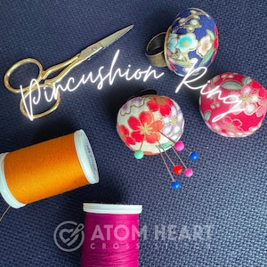 May include: Three colorful pincushion rings with floral patterns. The center ring has pins inserted and is surrounded by spools of thread. The text "Pincushion Ring" is written in white on a black background.
