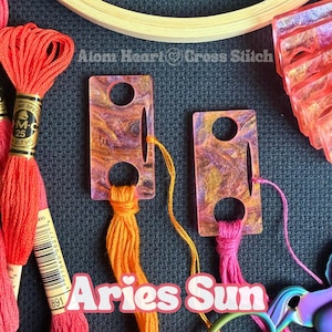 May include: Close-up of embroidery supplies, including orange and pink embroidery floss, two rectangular thread organizers, and a wooden hoop. The text "Aries Sun" is visible, along with "Atom Heart Cross Stitch".