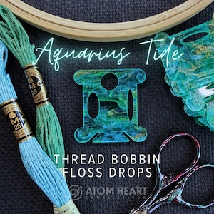 May include: An image featuring cross-stitch supplies. The text "Aquarius Tide" is displayed above a teal thread bobbin and floss drops. Two skeins of embroidery floss are visible, along with a pair of scissors and a wooden embroidery hoop.
