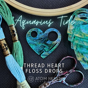 May include: A heart-shaped thread floss drop with a blue and green marbled design, next to embroidery floss and scissors. The text "Aquarius Tide" and "Thread Heart Floss Drops" are visible.