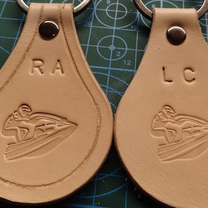 May include: Two beige leather keychains with silver key rings. Each keychain features a jet ski design and initials. One keychain has "RA" and the other has "LC" embossed on the top.