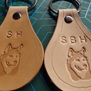 May include: Two tan leather keychains with metal rings. Each keychain has a dog silhouette and initials engraved on the front. The initials on the left keychain are "SH" and the initials on the right keychain are "SBH".
