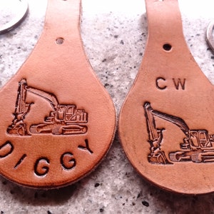 Könnte beinhalten: Zwei braune Leder-Schlüsselanhänger mit einem Bagger-Design und personalisierten Namen. Der Schlüsselanhänger links trägt die Aufschrift "DIGGY" und der Schlüsselanhänger rechts "CW".