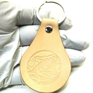 Pode incluir: Um chaveiro de couro bege com um chaveiro de prata. O chaveiro tem um design circular com um animal de desenho animado dentro.