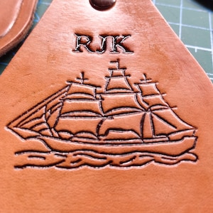 May include: A brown leather keychain with the initials "RJK" embossed on it. A detailed image of a sailing ship is also embossed on the keychain.