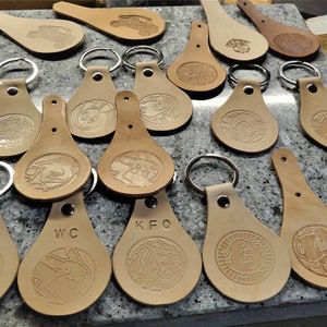 Könnte beinhalten: Eine Sammlung von 14 Schlüsselanhängern aus braunem Leder mit gravierten Designs und Initialen. Jeder Schlüsselanhänger hat einen silbernen Metallring.