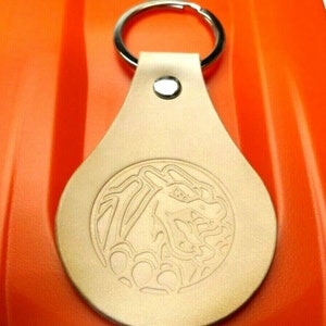 May include: A tan leather keychain with a silver key ring. The keychain has a circular design of a dragon with its mouth open, embossed on the front.