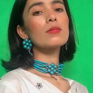 May include: A woman with dark hair wearing a white shirt with black floral embroidery and a turquoise beaded necklace and earrings. The background is a solid green.