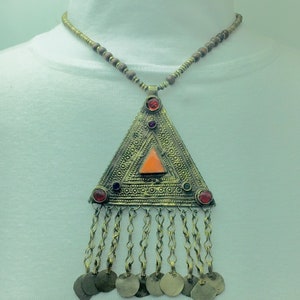 Puede incluir: Un collar con un colgante de triángulo dorado adornado con piedras preciosas rojas, naranjas y azules. El colgante tiene una franja de pequeñas cadenas y monedas doradas.