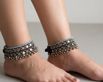 May include: A pair of anklets featuring multiple rows of small, round, metallic silver-toned discs and tiny dangling bells. The upper edge of each anklet is accented with a single row of light blue beads, all set against a dark fabric base.
