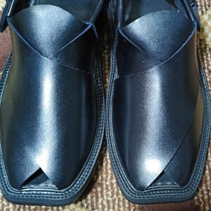 May include: A pair of black leather shoes with a pointed toe and a decorative stitch around the edge. The shoes have a unique design with a strap that crosses over the top of the foot.