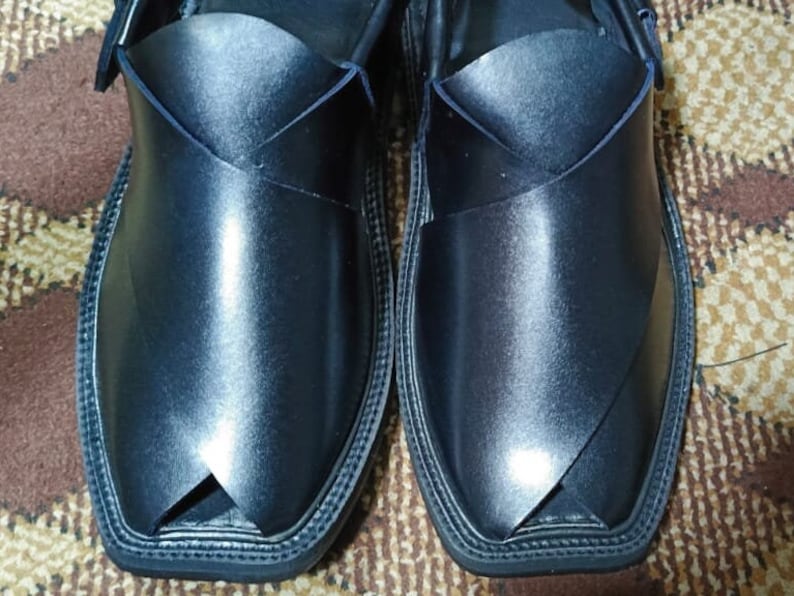 May include: A pair of black leather shoes with a pointed toe and a decorative stitch around the edge. The shoes have a unique design with a strap that crosses over the top of the foot.