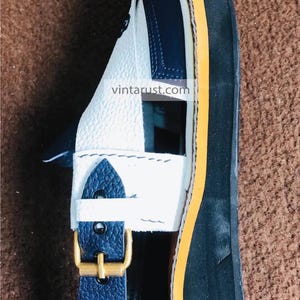 Pu&ograve; includere: Sandali in pelle bianca e blu con bordino giallo e chiusura a fibbia. I sandali hanno una suola nera spessa. I sandali sono fatti a mano e sono in stile vintage.