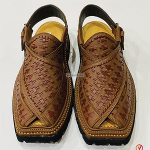 May include: A pair of brown leather Peshawari sandals with a woven design. The sandals have a strap around the ankle and a black sole. The leather is a medium brown color with darker brown accents. The sandals are handmade.