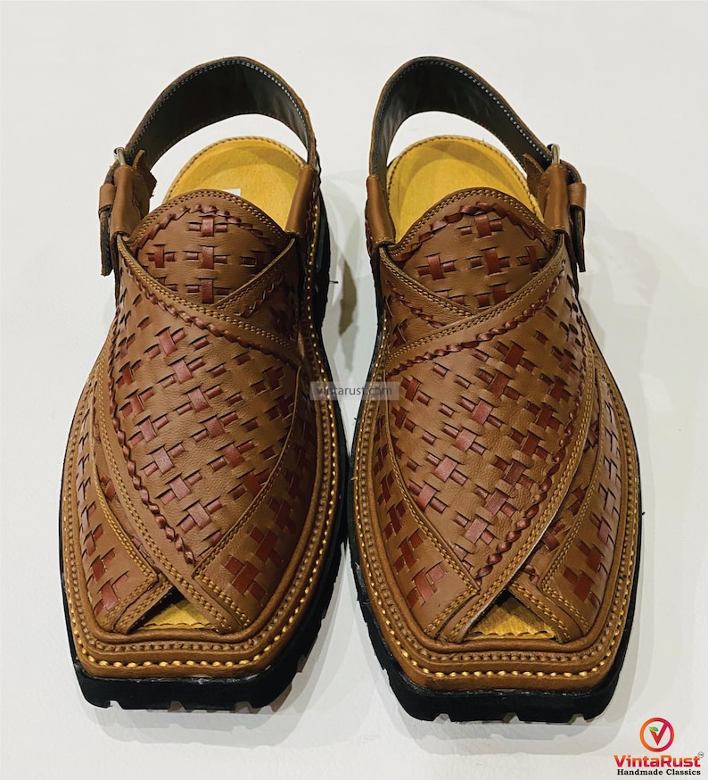 May include: A pair of brown leather Peshawari sandals with a woven design. The sandals have a strap around the ankle and a black sole. The leather is a medium brown color with darker brown accents. The sandals are handmade.