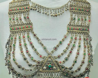 May include: A silver and gold necklace with multiple strands of chains and beads. The necklace features a triangular pendant with green and red stones. The necklace is a traditional style of jewellery.