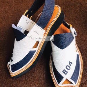 Pu&ograve; includere: Un paio di sandali in pelle bianca e blu con suola nera. I sandali hanno un cinturino che va intorno alla caviglia e una chiusura a fibbia. I sandali hanno il numero "804" scritto su di loro.