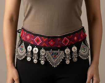 May include: A wide fabric belt featuring a red and black geometric pattern with a black border. The belt is adorned with multiple silver-toned metal charms, including circular discs and a large central triangular pendant with colorful accents.