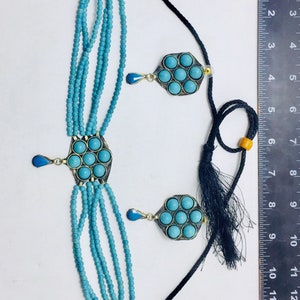 May include: A turquoise blue beaded necklace with two pendants featuring turquoise stones set in silver. The necklace is on a white background with a ruler for scale.
