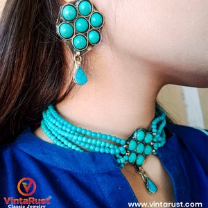 May include: A turquoise blue choker necklace with multiple strands of beads and a pendant featuring a flower design with turquoise stones. The pendant has a teardrop-shaped dangle. Matching turquoise earrings with a flower design and teardrop dangle.