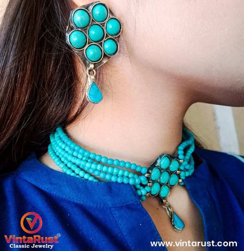 May include: A turquoise blue choker necklace with multiple strands of beads and a pendant featuring a flower design with turquoise stones. The pendant has a teardrop-shaped dangle. Matching turquoise earrings with a flower design and teardrop dangle.