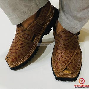 May include: Close-up of two brown leather Peshawari sandals with a woven pattern and black soles. The sandals have a strap across the top and are worn by someone wearing light gray pants. The sandals are handmade classics.
