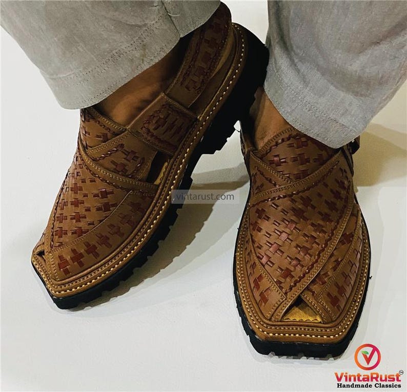May include: Close-up of two brown leather Peshawari sandals with a woven pattern and black soles. The sandals have a strap across the top and are worn by someone wearing light gray pants. The sandals are handmade classics.