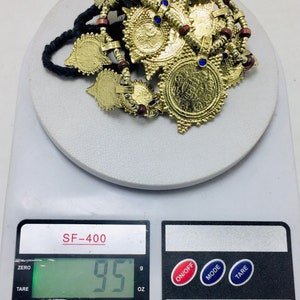 May include: A gold-coloured necklace with multiple circular pendants and red and blue beads. The necklace is on a digital scale that reads 95 grams.