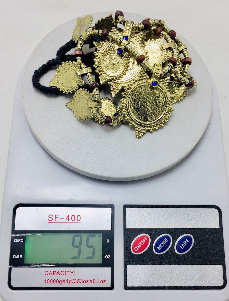 May include: A gold-coloured necklace with multiple circular pendants and red and blue beads. The necklace is on a digital scale that reads 95 grams.