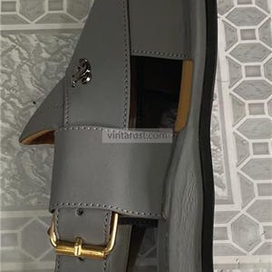 K&ouml;nnte beinhalten: Graue Ledersandale mit goldener Schnalle und dekorativem silbernen Emblem. Die Sandale hat eine schwarze Sohle und einen Riemen &uuml;ber dem Fu&szlig;. Die Sandale ist Gr&ouml;&szlig;e 43 (EU).