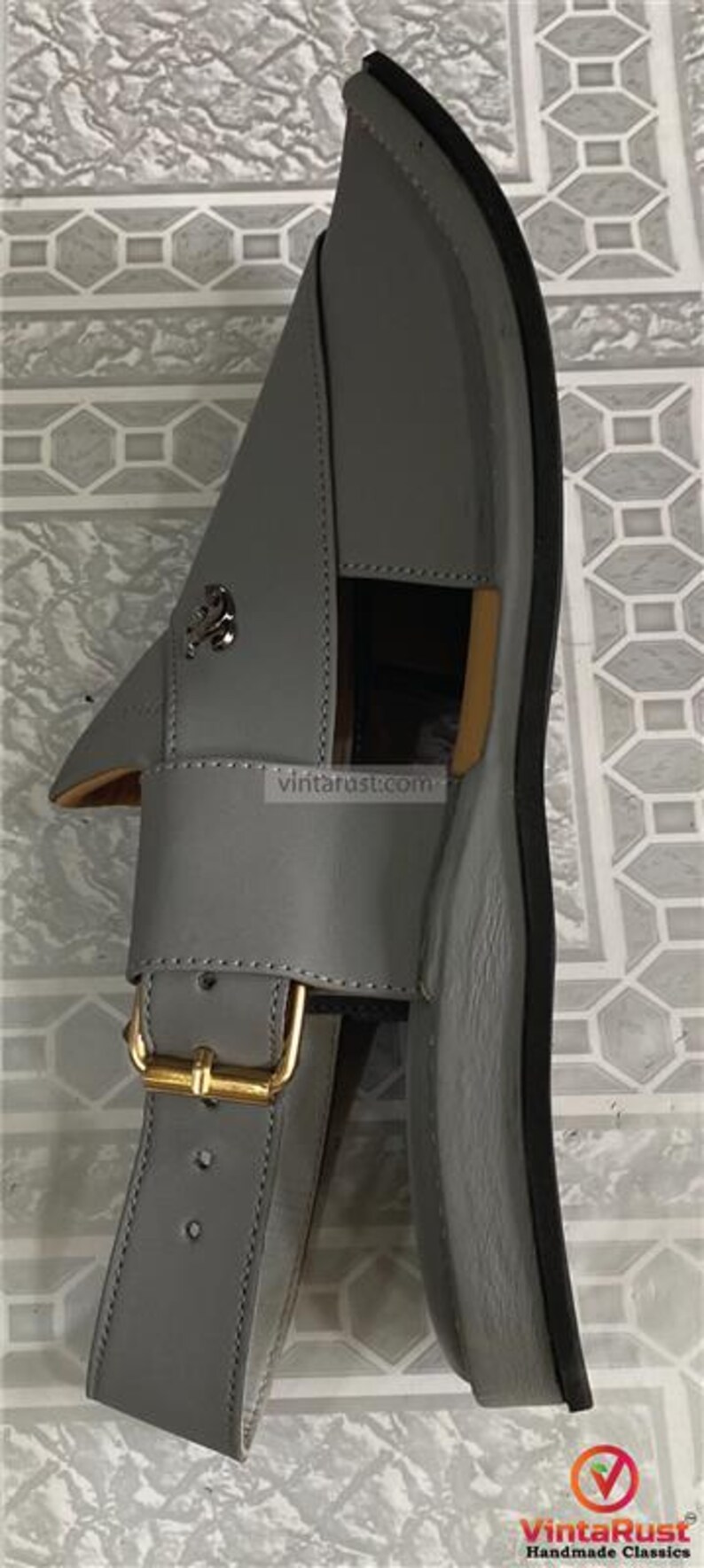 K&ouml;nnte beinhalten: Graue Ledersandale mit goldener Schnalle und dekorativem silbernen Emblem. Die Sandale hat eine schwarze Sohle und einen Riemen &uuml;ber dem Fu&szlig;. Die Sandale ist Gr&ouml;&szlig;e 43 (EU).