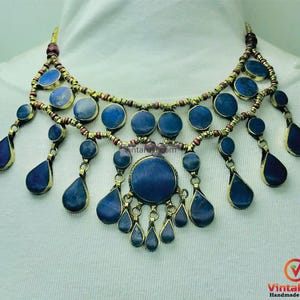 May include: A gold necklace with a layered design featuring blue lapis lazuli stones. The necklace has a central pendant with a larger lapis lazuli stone and smaller stones hanging from it.