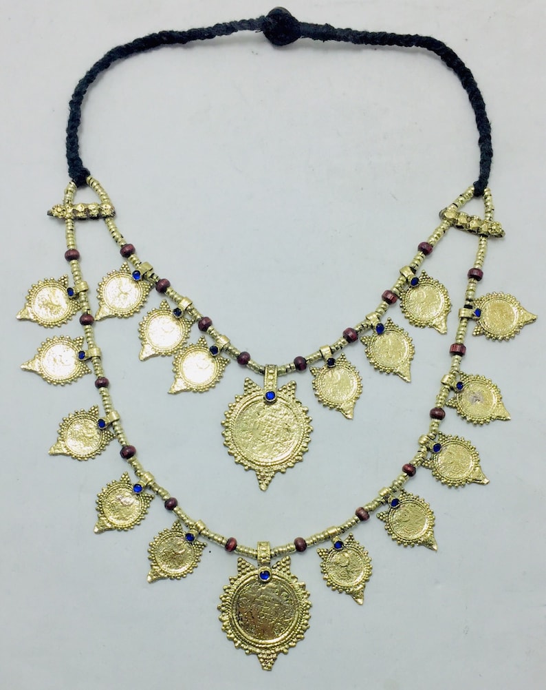 May include: A gold necklace with a double strand of brown beads and gold coin-shaped pendants. The pendants have blue gemstones in the centre. The necklace is tied with a black cord.