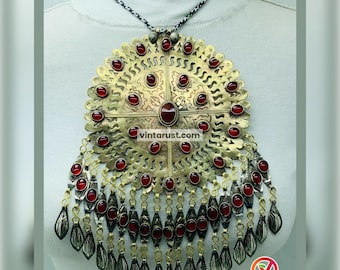 May include: A large, round, gold-toned pendant necklace with a intricate design and red gemstone accents. The pendant features a cross shape with a central red gemstone and a fringe of smaller red gemstones.